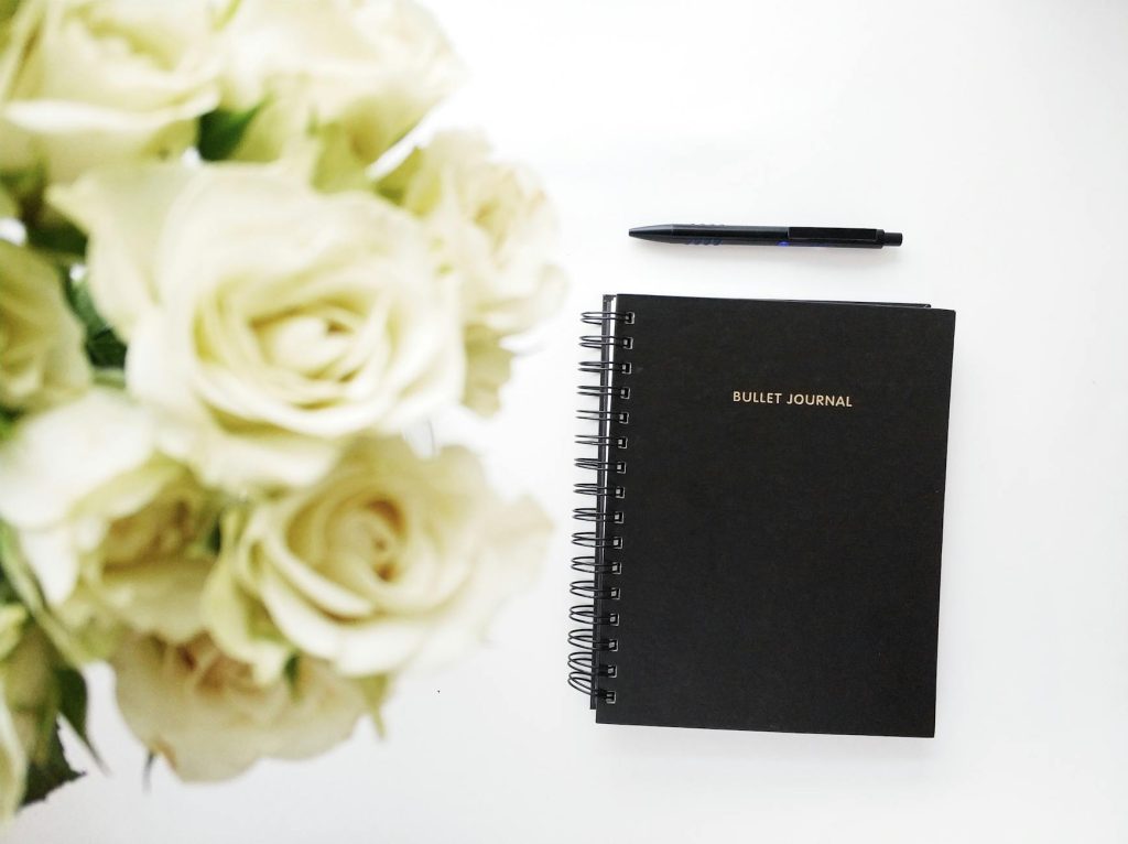Flat lay of a black bullet journal with a bouquet of white roses, creating a stylish and romantic workspace arrangement.
