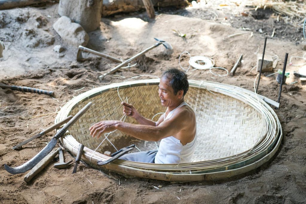 An Asian man skillfully weaving a large bamboo basket outdoors, surrounded by tools and natural materials.