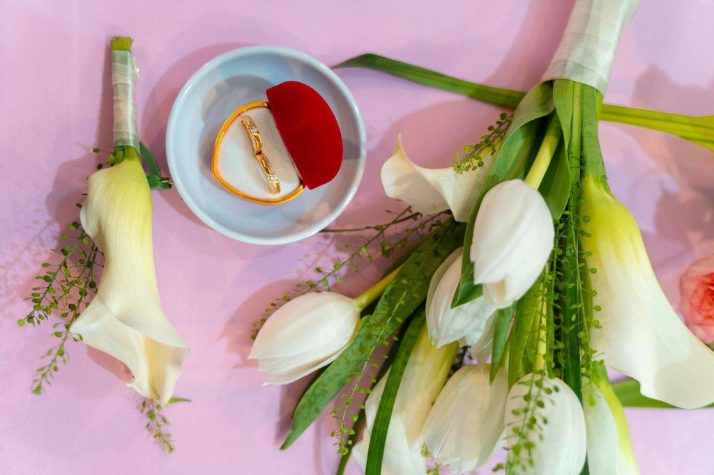 Flat lay of wedding rings in a decorative box with white tulips and calla lilies on pink background.