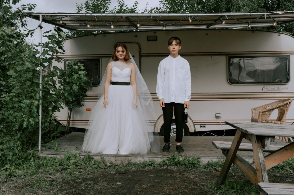 A bride and groom pose with a vintage caravan in the background, highlighting a rustic wedding theme.