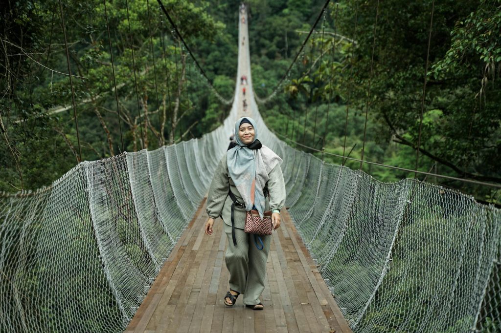 A woman in hijab walks confidently on a scenic suspension bridge in Sukabumi, Indonesia.