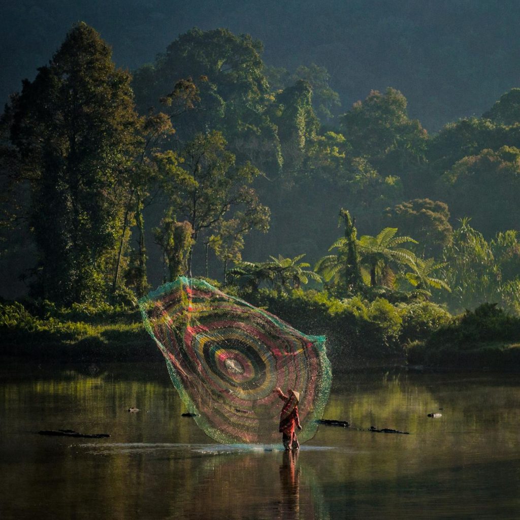 Fisherman casting colorful net on serene lake amidst dense Sukabumi forest landscape.