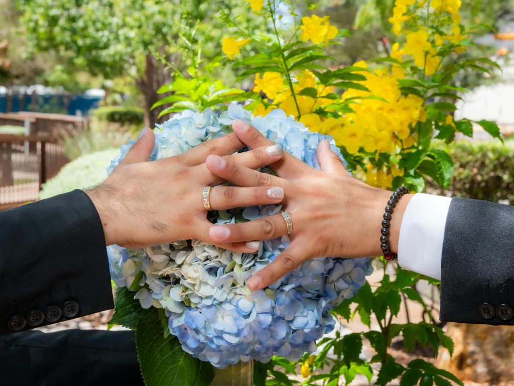 Close-up of wedding rings on hands over a blue hydrangea bouquet outdoors.