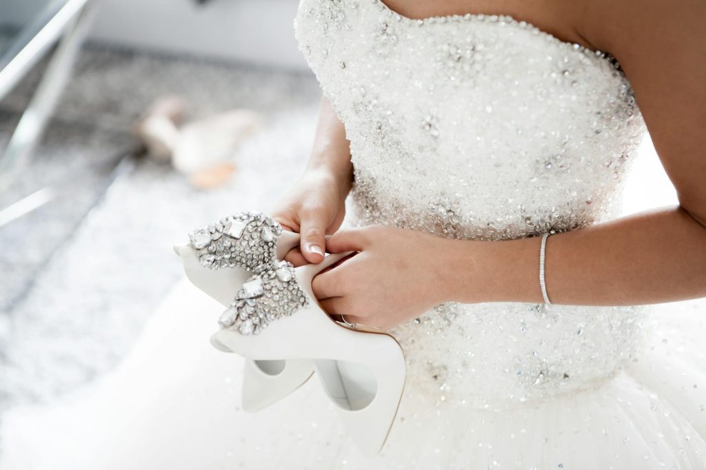 Bride in a dazzling wedding dress holding embellished high heels.