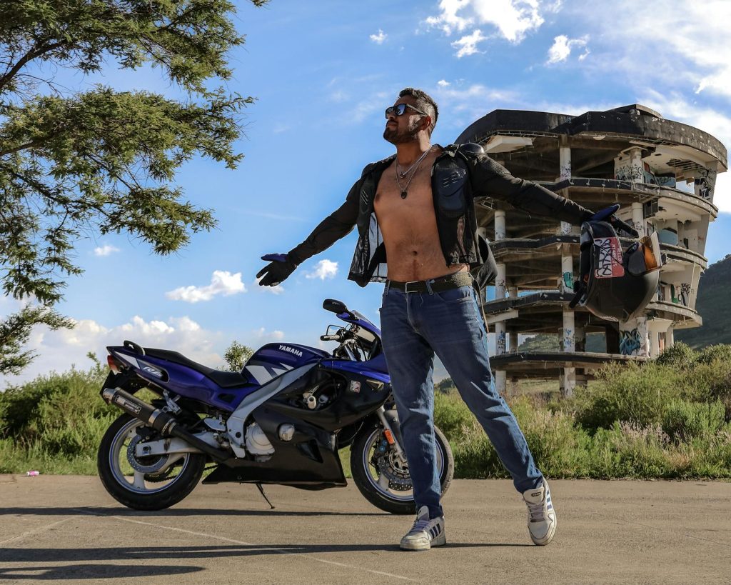 Confident man with motorcycle jacket posing outside an abandoned building near nature.