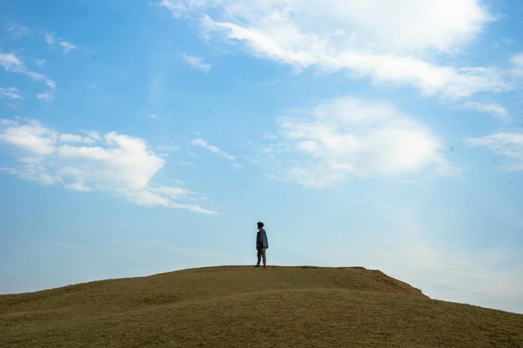 A lone traveler stands on a grassy hilltop under a vast blue sky, capturing a serene moment in Ghale Gaun, Nepal.