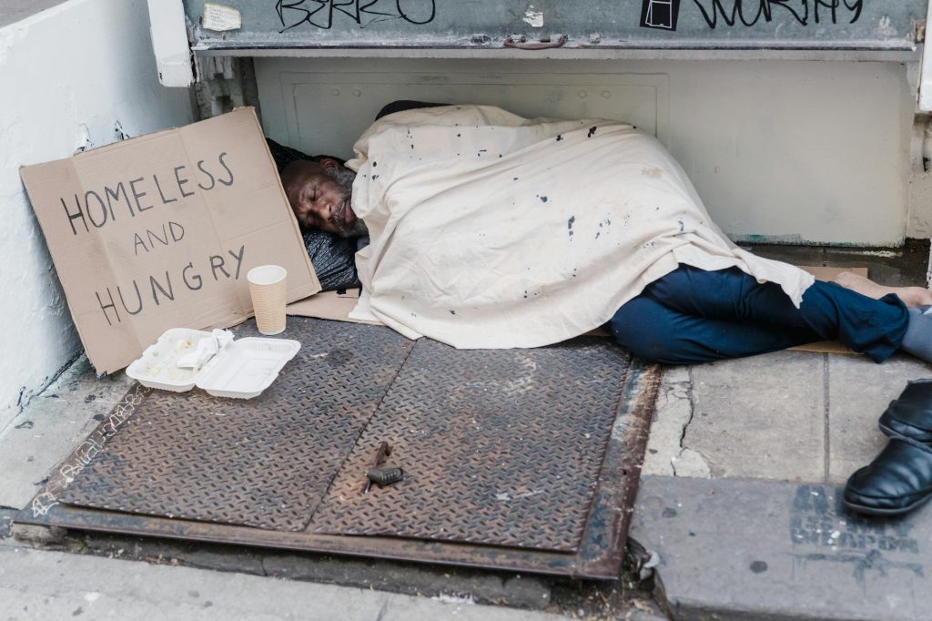 Street scene depicting a homeless man sleeping with a placard reading 'Homeless and Hungry'.