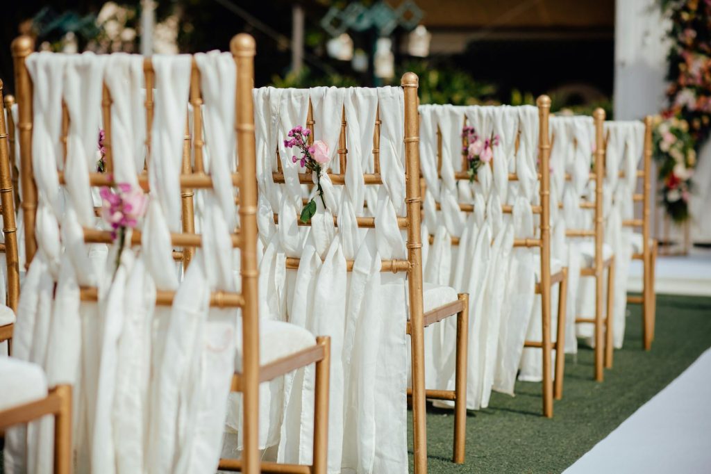 Row of elegant wooden chairs decorated with white fabric and pink flowers for an outdoor wedding ceremony.