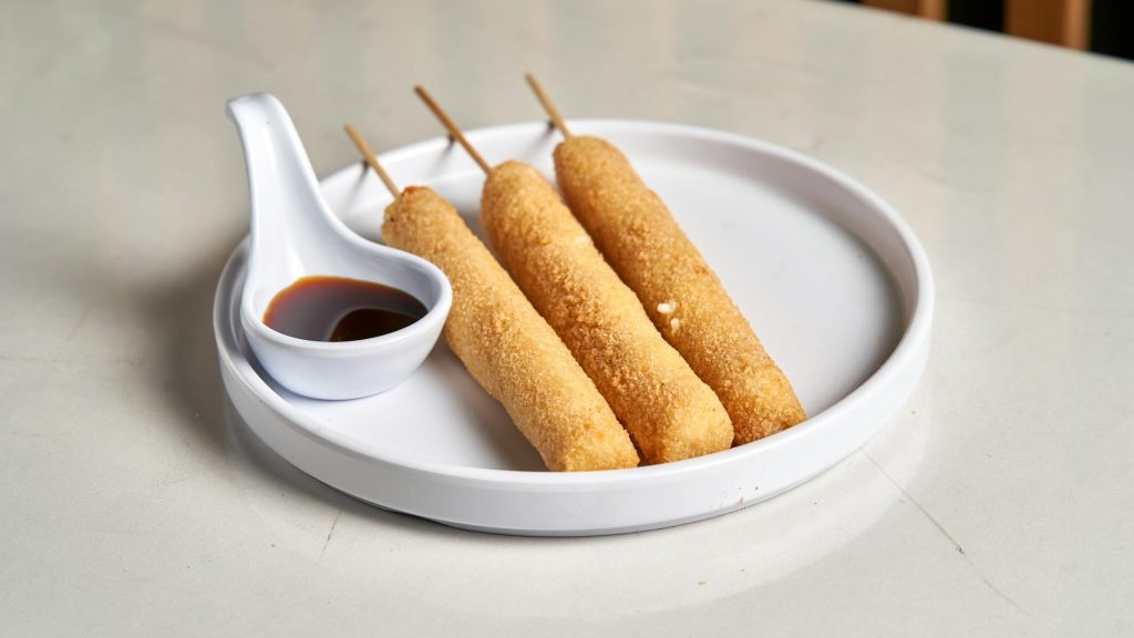 Three crispy skewered snacks served with dipping sauce on a white plate.
