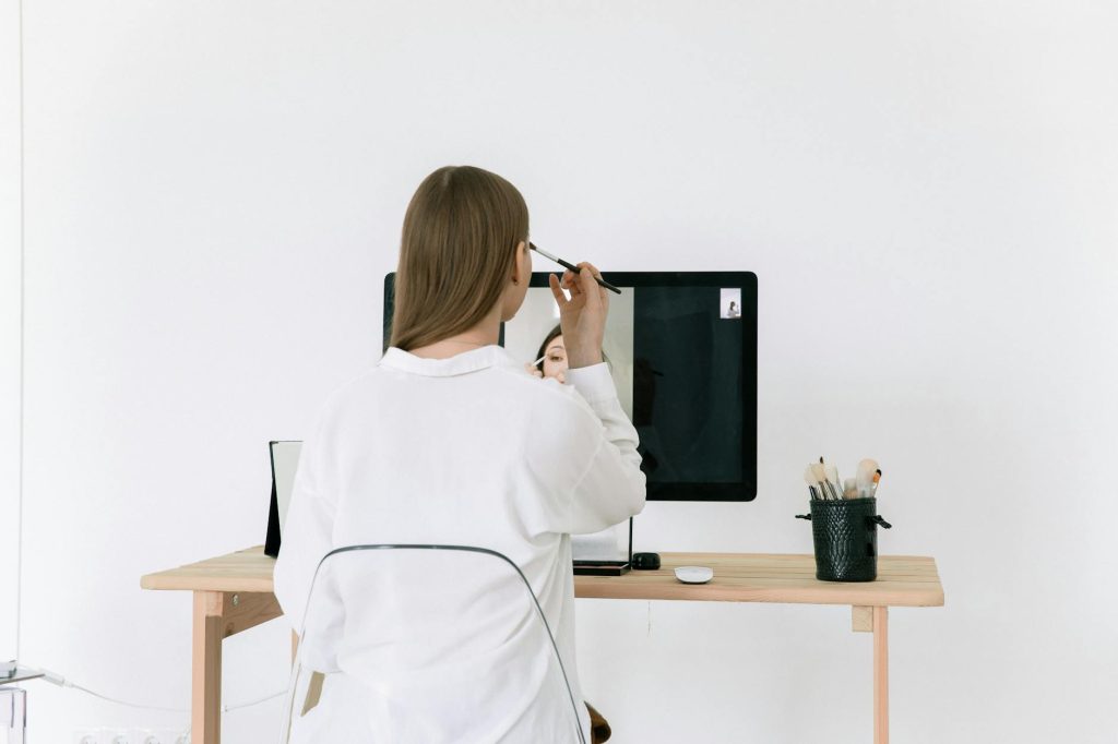 A woman applies makeup at her desk using a computer screen as a mirror.
