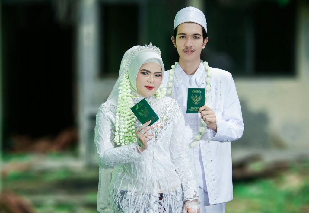 A joyful Indonesian couple celebrating their Muslim wedding in traditional attire, holding marriage certificates.