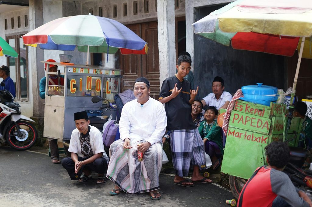 Group of men wearing traditional attire relaxing at a bustling street food market with colorful stalls.