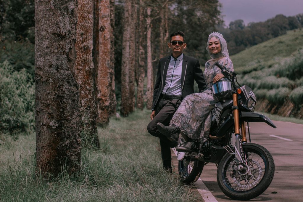 A fashionable couple posing with a motorcycle in a scenic forest setting, capturing happy expressions.