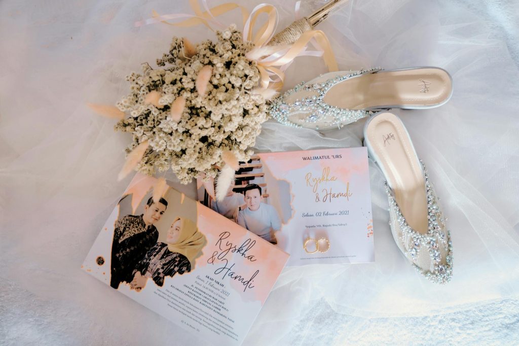 Flat lay of wedding invitation with shoes and floral bouquet on a white sheet.